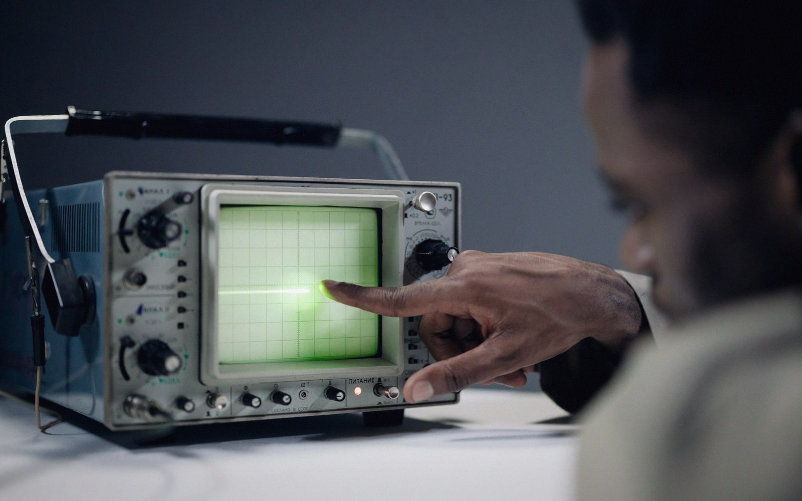 A person points a finger at a glowing green line on an oscilloscope screen